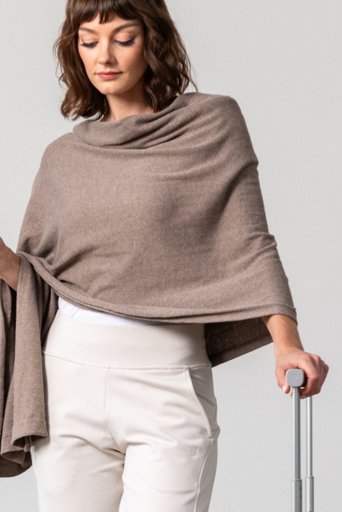 Women shown wearing a dark beige scarf draped over her shoulders with one hand on her luggage handle